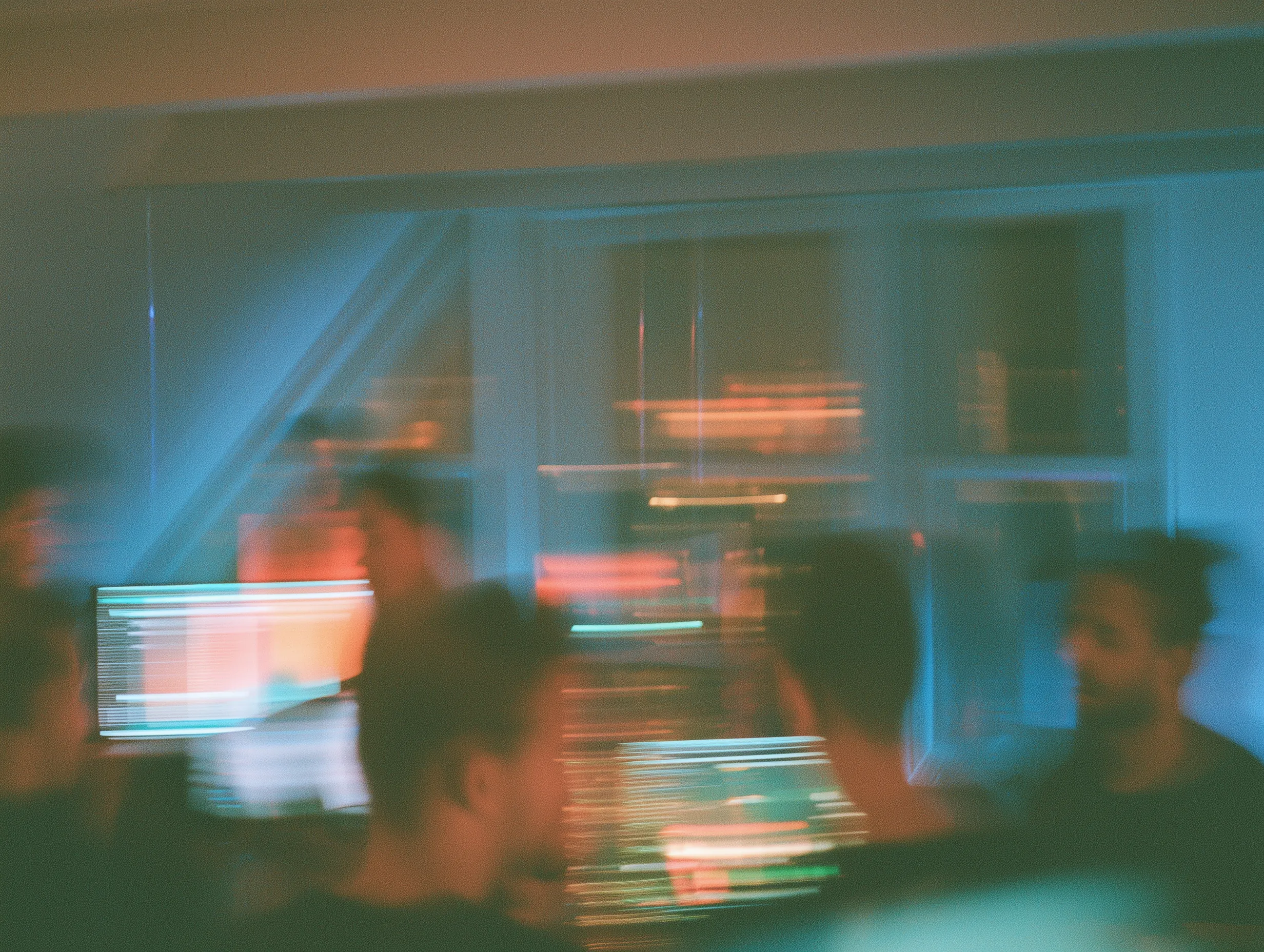 A motion-blurred view of colleagues collaborating around glowing computer screens in a dark workspace.